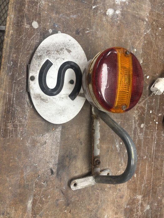 Gammal S-skylt och rund baklykta i behov av renovering, liggande på ett slitet träbord i en verkstad. Skruvar och fästen synliga.
