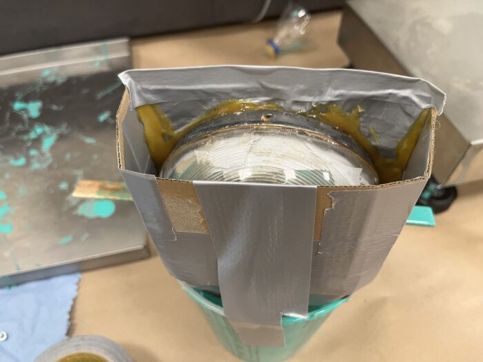 Gjutform av kartong och silvertejp kring en baklykta, fylld med silikon för att skapa en form. Verktyg och kladdigt arbetsområde syns i bakgrunden.