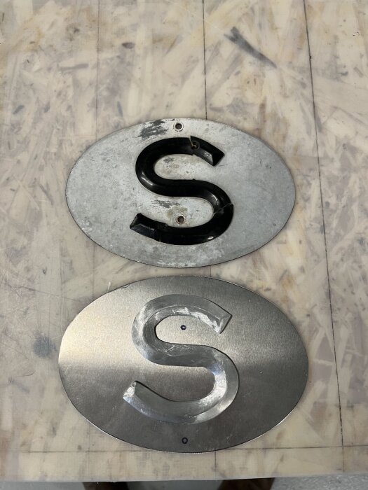 Två ovala S-skyltar i metall, en gammal och sliten med svart plast-S, den andra ny och blank med inristat S.