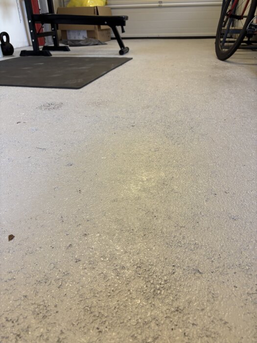 Betonggolv i garage nymålat med tvåkomponents epoxyfärg, omgiven av träningsutrustning och cykelhjul.
