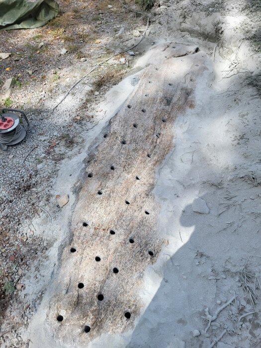 Bergplatta med många borrade hål för klyvning; omgivning av grus och jord, synligt förlängningskabel på marken bredvid.