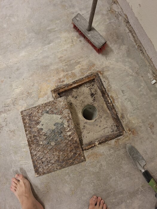 Gammal plåtlucka upplyft, exponerar ett runt renshål i golvet; kvast och verktyg syns på betonggolvet, nakna fötter nedtill.