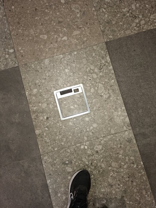 Klinkergolv med inbyggd metallucka och en fot i sneakers syns i bilden.