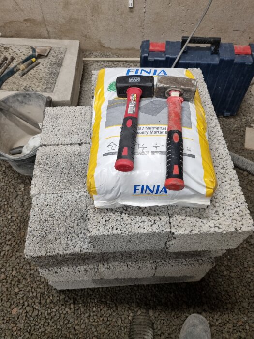 Verktyg, murbrukpåse och betongblock på golvet i en byggmiljö.