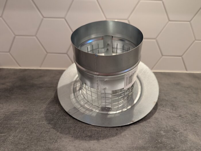 Metallisk takhuv med galler, stående på en yta framför vit kakelvägg. Rör i ovankant, skarv synlig.