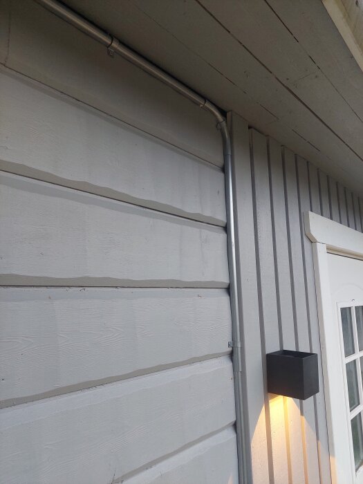 Kabeldragning längs väggen vid ett hus, med eluttag vid takfoten för infravärme och ljusslinga; en svart utomhuslampa är tänd.