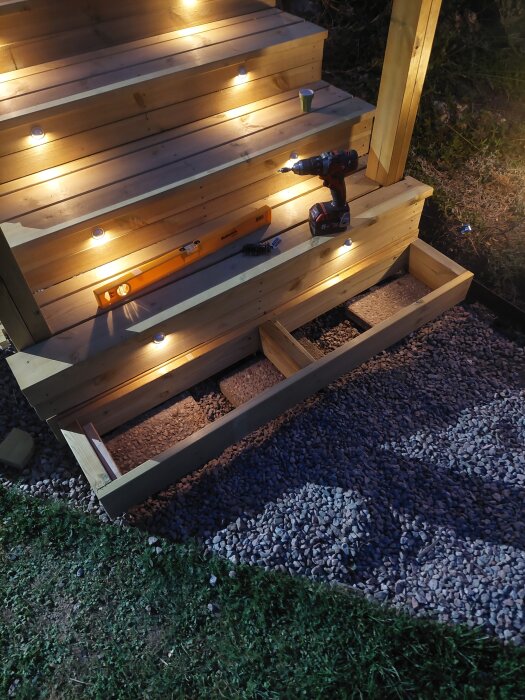 Trätrappa utomhus med inbyggda LED-lampor, en skruvdragare, ett vattenpass och grus vid trappens bas, fotograferad i skymning.