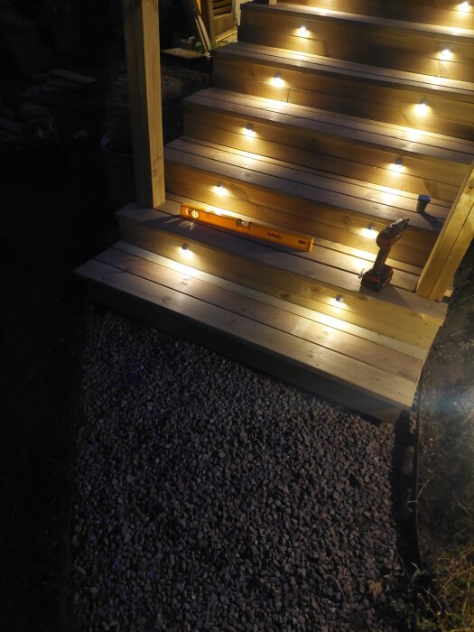 Trätrappa med monterad belysning i stegen, grusgång vid nedersta steget, skruvdragare och vattenpass synliga.