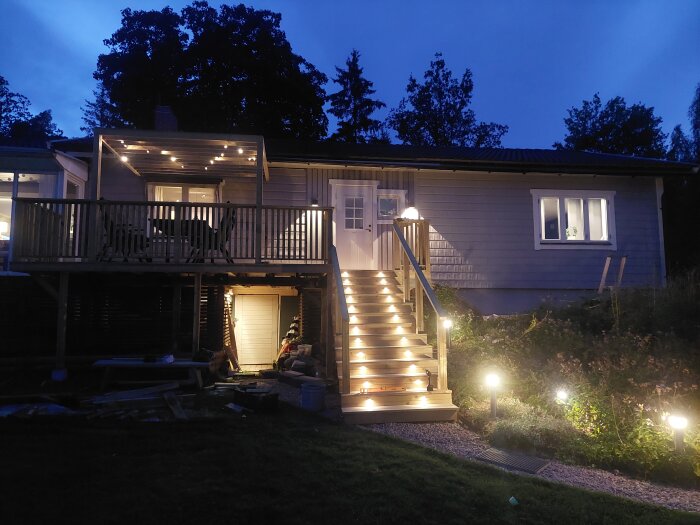 Upplyst veranda och trappa på ett hus i skymningen, omgiven av buskar och med en ljusslinga på pergolan.