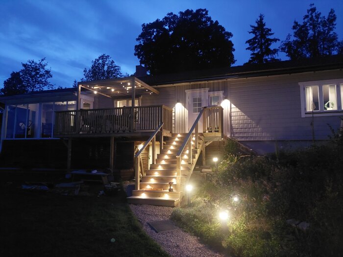 Upplyst trappa och altan med ljusslinga under en klar kvällshimmel. Trappsteg leder till en dörr på väggen av ett hus.