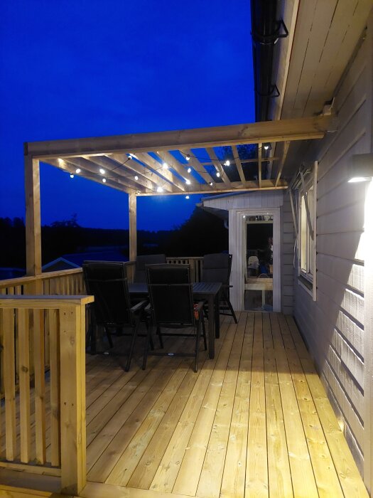 Träterrass med pergola dekorerad med ljusslinga, utemöbler och kvällsbelysning vid husfasad.