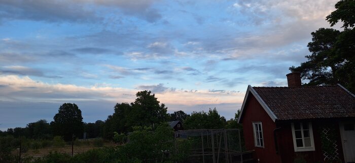 Rött hus med tegeltak i förgrunden, omgiven av grönska och träd, med en blå och molnig himmel i bakgrunden.