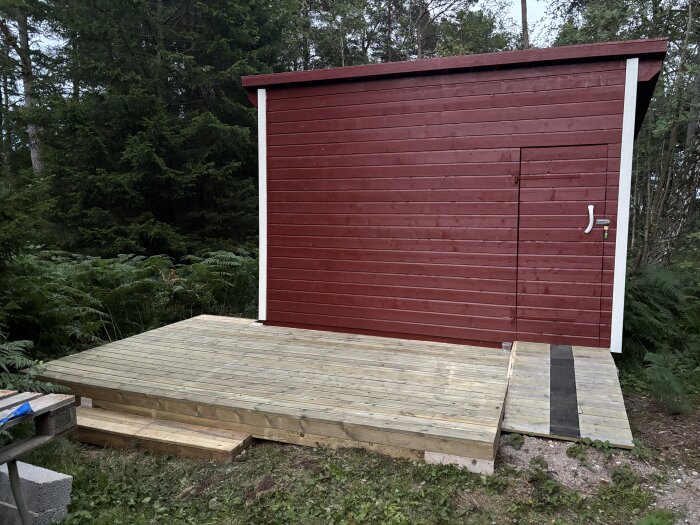 Rött förråd med litet trädäck framför, omgivet av grönskande skog.