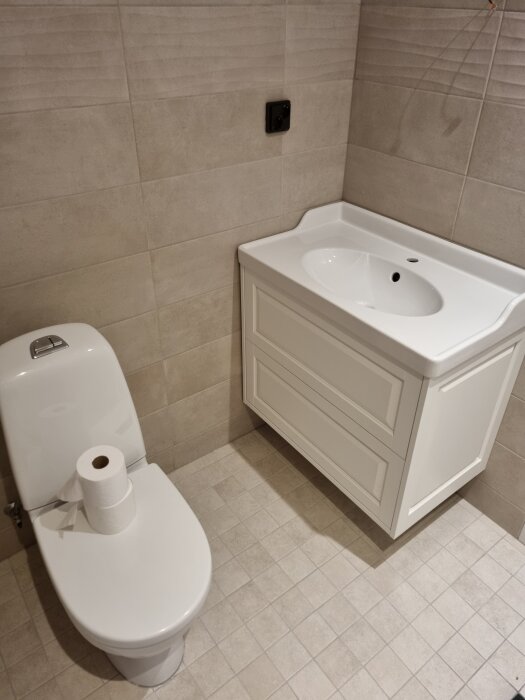 Modernt badrum med kaklade väggar, toalett och vita badrumsskåp. Toalettpapper placerat ovanpå toalettstolen.