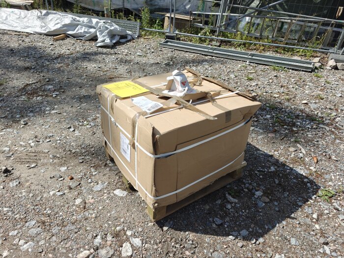 Pall med paket inslaget i kartong och plasticsnören utomhus på grusig mark med byggställningar i bakgrunden.