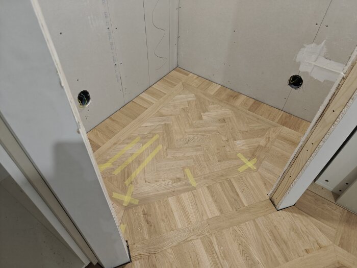 Parkettgolv med fiskbensmönster i rum under renovering, gipsväggar med målade hål för elinstallation och tejp som markerar områden på golvet.
