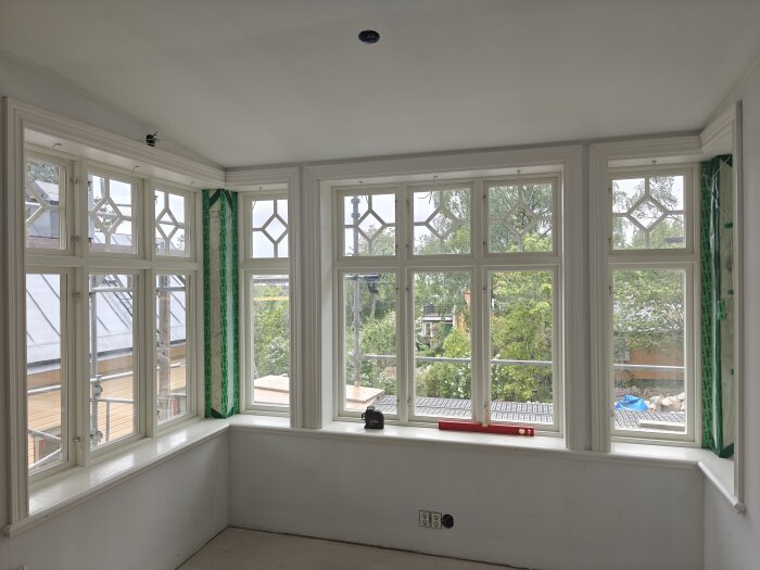 Glasveranda med stora spröjsade fönster som släpper in ljus, utanför syns träd och byggställning.