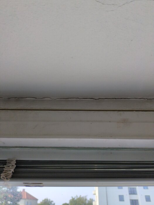 Sprickor i vitmålad betong ovanför ett fönster, synliga mellan fönsterkarm och vägg.