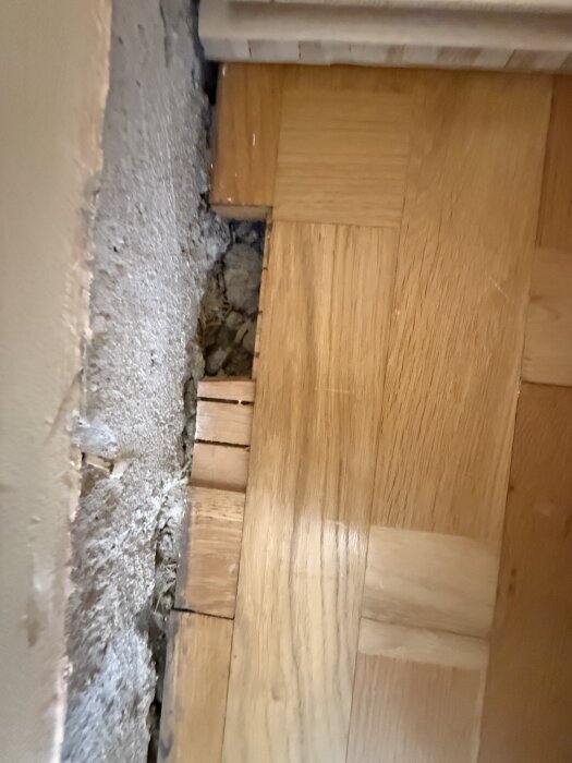 Närbild av trägolv under borttagen parkett, visar träets ådring och färg, med en öppning mot en vägg i bakgrunden.