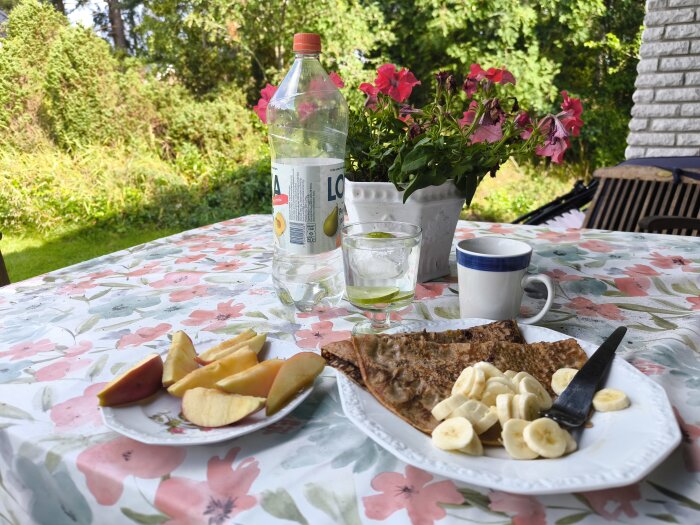 Pannkakor med bananmusli, skivad banan, Àppelklyftor, en flaska Loka, kaffe och blommor pÄ blommig duk.