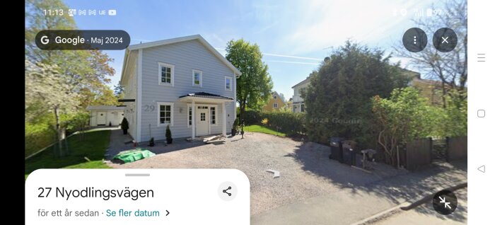 Tvåvåningshus i vitt med brunt tak och vita fönster, omgiven av träd och buskar, på en grusad uppfart.