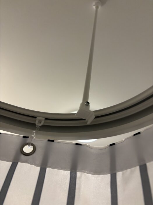 Duschspiralstång med trasig monteringsdel mot taket. Strimmigt duschdraperi hänger från stången.