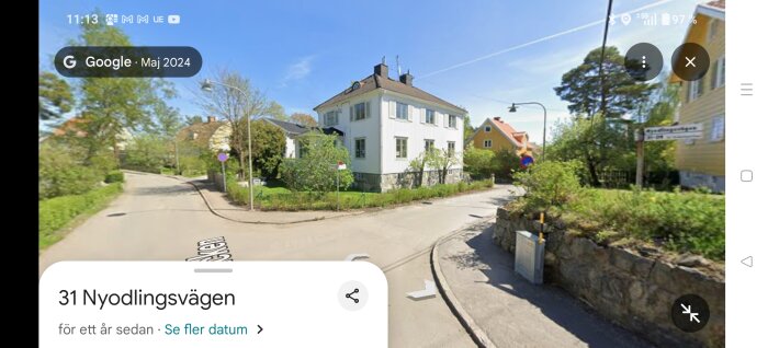 Vita tvåvåningshus i hörn av Nyodlingsvägen, grönskande omgivning, tydlig vägskylt, blå himmel i bakgrunden.