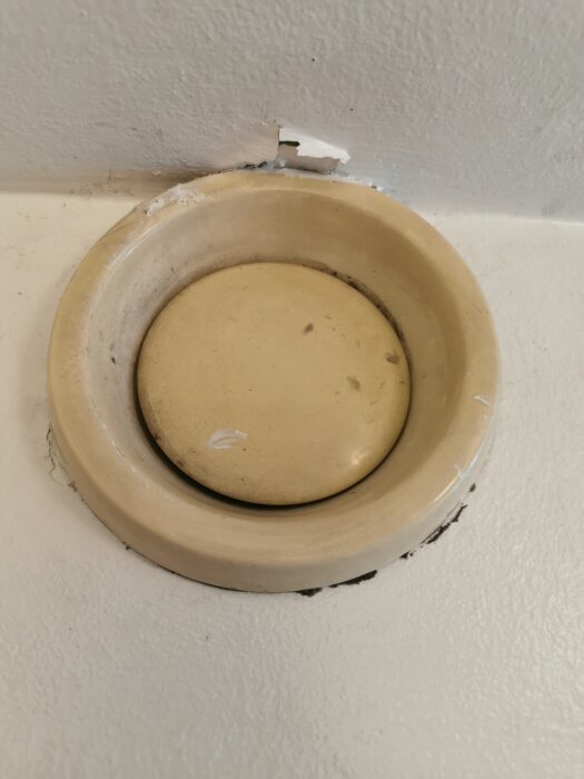 Gammalt frånluftsdon i beige färg monterat i badrumsvägg, med sprickor och väggsläpp runtomkring.
