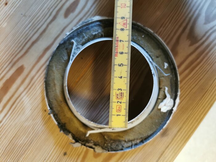 Gammalt frånluftsdon med smutsig kant, placerat på ett trägolv. En tumstock mäter innerdiametern.