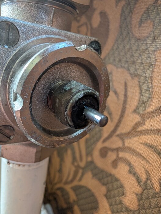 Närbild på en metallpackbox med en liten läckage, monterad på ett rör intill en vägghängd tapet med brunt mönster.