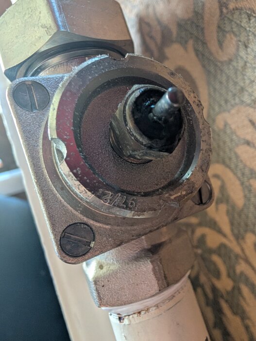 Närbild av en läckande packbox på ett rörsystem med synlig pigg och koppling, installerad mot en mönstrad väggbakgrund.