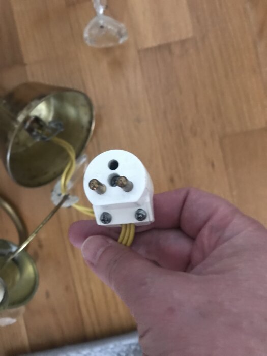 Hand som håller i en vit elkontakt med gul kabel, lampdelar syns i bakgrunden på trägolv.