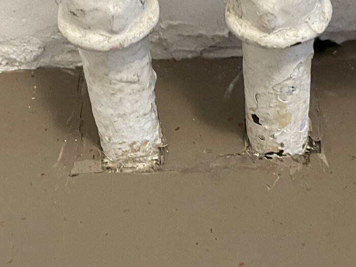 Linoleummatta runt två rör med synliga skarvar och sprickor där mattan inte sitter ordentligt fast vid kanterna.