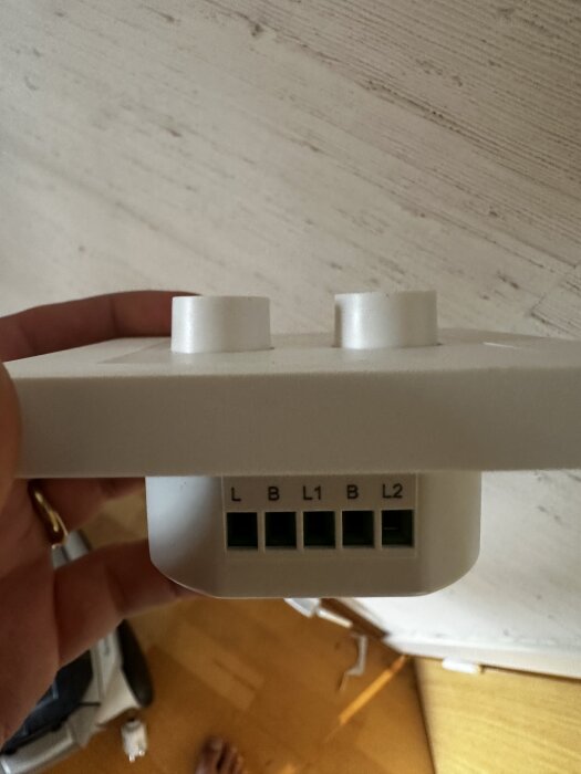 Kronströmbrytare med anslutningar märkta L, B, L1, B, L2, hålls av en hand mot en vit väg.