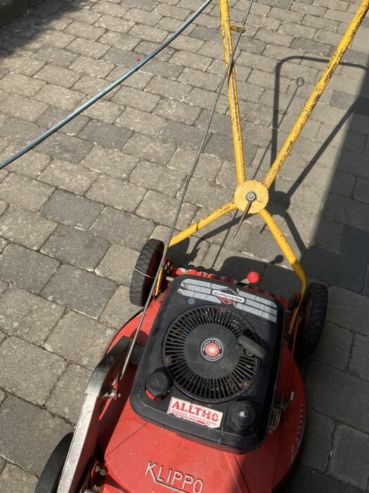 Röd gräsklippare av märket Klippo med gul handtag och vajrar, stående på stenlagd mark, fotograferad uppifrån.