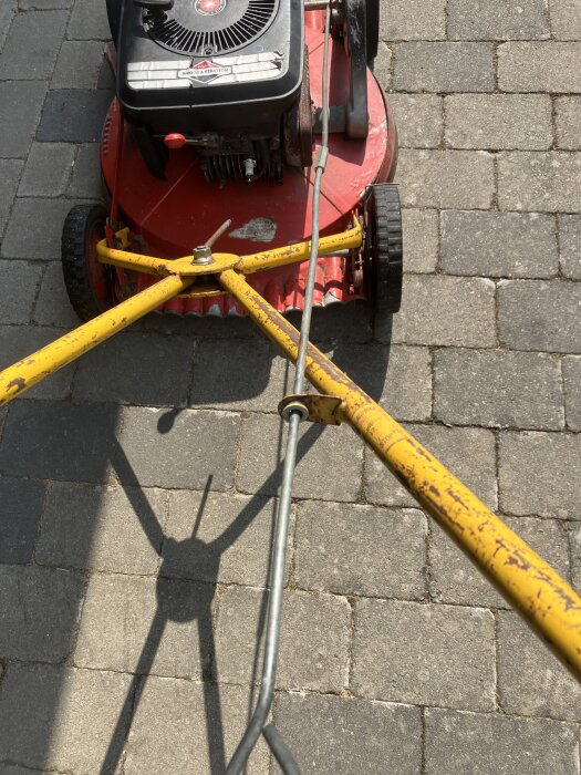 Röd gräsklippare med gul stång och motor ovanpå, stående på stenbelagd yta, del av diskussion om maskinens självgångsfunktion och eventuella saknade delar.