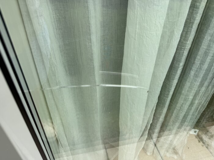 Spricka i mellanglaset på ett treglasfönster, synlig mot en bakgrund av ljusa gardiner. Bilden visar problem med fönstret.