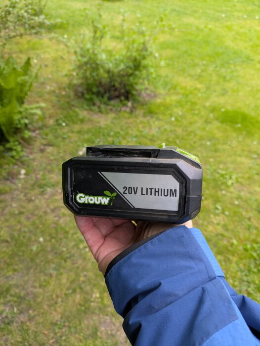 Hand som håller i ett 20V litiumbatteri från märket Grouw, fotograferat utomhus på en grön gräsmatta.