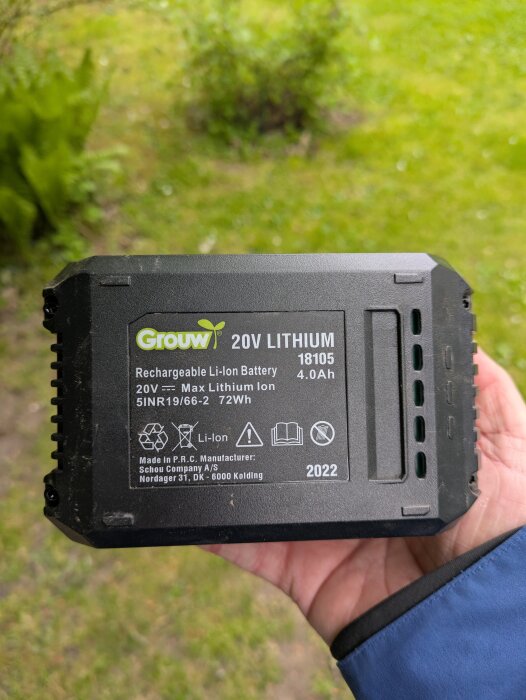 Uppladdningsbart 20V litiumjonbatteri (Grouw) hålls i en hand, med grön bakgrund av gräs och buskar.
