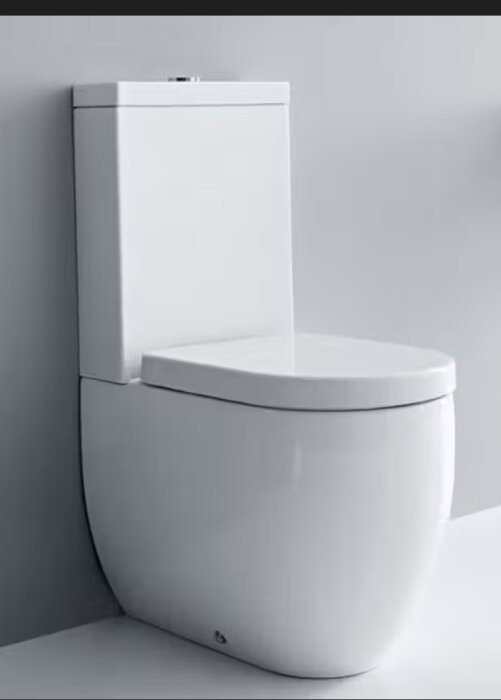 Vit golvstående toalett från Lavabo Flo mot ljusgrå vägg, med slät front och cistern. Inget sidouttag för vattenanslutning syns.