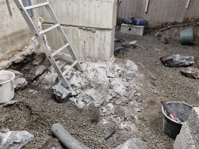 Byggarbetsplats med grus, stenar och en stege vid en betongvägg; verktyg och hinkar syns intill en kabel på marken.