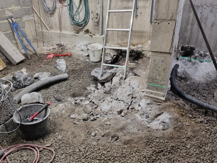 Byggarbetsplats med metallstege, en hink, dräneringsrör i plast och krossad betong kring en betongpelare, inomhusväggar i bakgrunden.