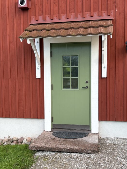 Grönmålad teakdörr med vita karmar på ett rött hus, med brunt takutsprång ovanför. Stenlagd entré med svart dörrmatta.