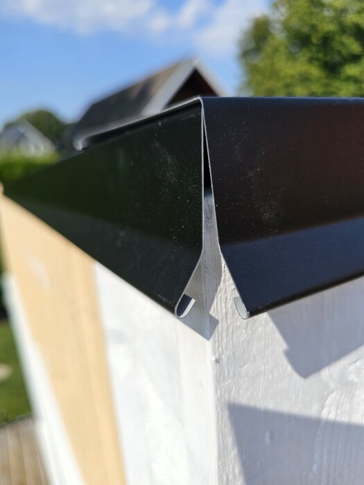 Svart plåtgavelbeslag monterat på en husfasad utan vindskivor, med provisoriskt lagt plåt visar ett hörn med blå himmel i bakgrunden.