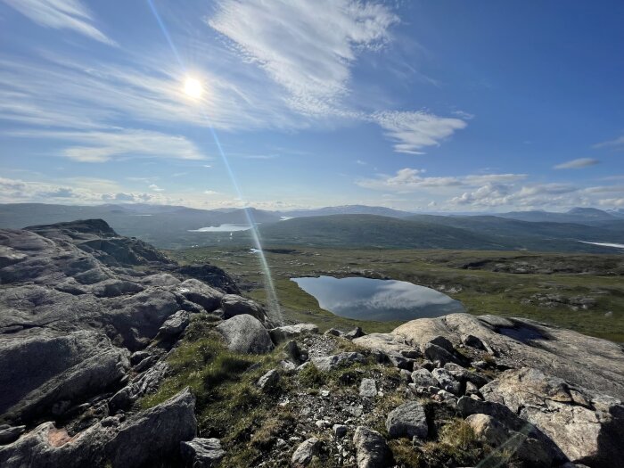 Solbelyst fjällandskap med klippor i förgrunden och en sjö i dalen, blå himmel med tunna moln över horisonten.