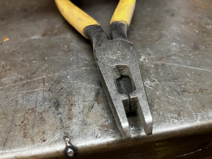 Specialiserad tång med gul handtag och rundad spets, som lämnar mellanrum när den är stängd, placerad på en metallisk yta.