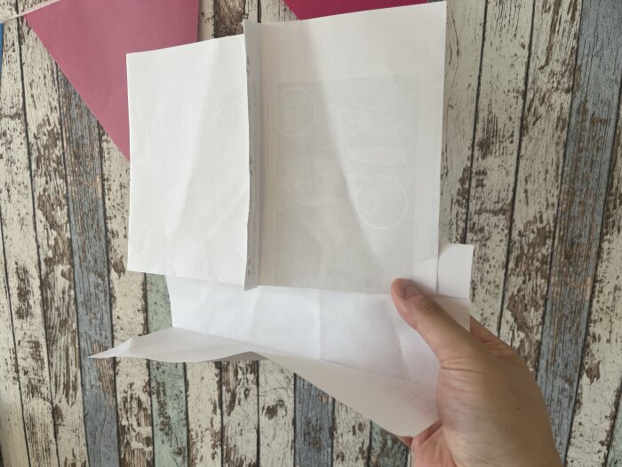 Hand håller en modell av papper med vikta sektioner, placerad mot en trätexturerad bakgrund.