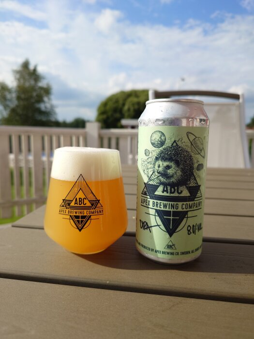 Aluminiumförpackning av Apex Brewing Company DIPA bredvid ett glas med skummande öl på ett bord utomhus en solig dag.