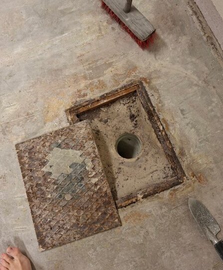 Öppen golvlucka med synligt avloppsrör i ett betonggolv, omgivet av verktyg och en fot; oklart omgivande rum.