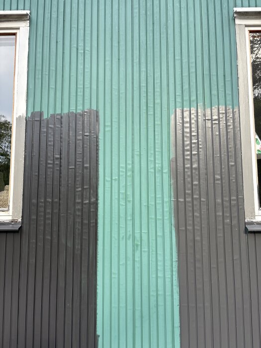 Grönmålad husfasad med blåsor och ny mörkare färg provmålad på vissa delar, inklusive två fönster.